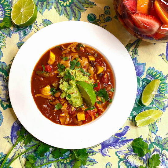 Vegan chili on a yellow, blue, and green tablecloth with a pitcher of sangria mocktail